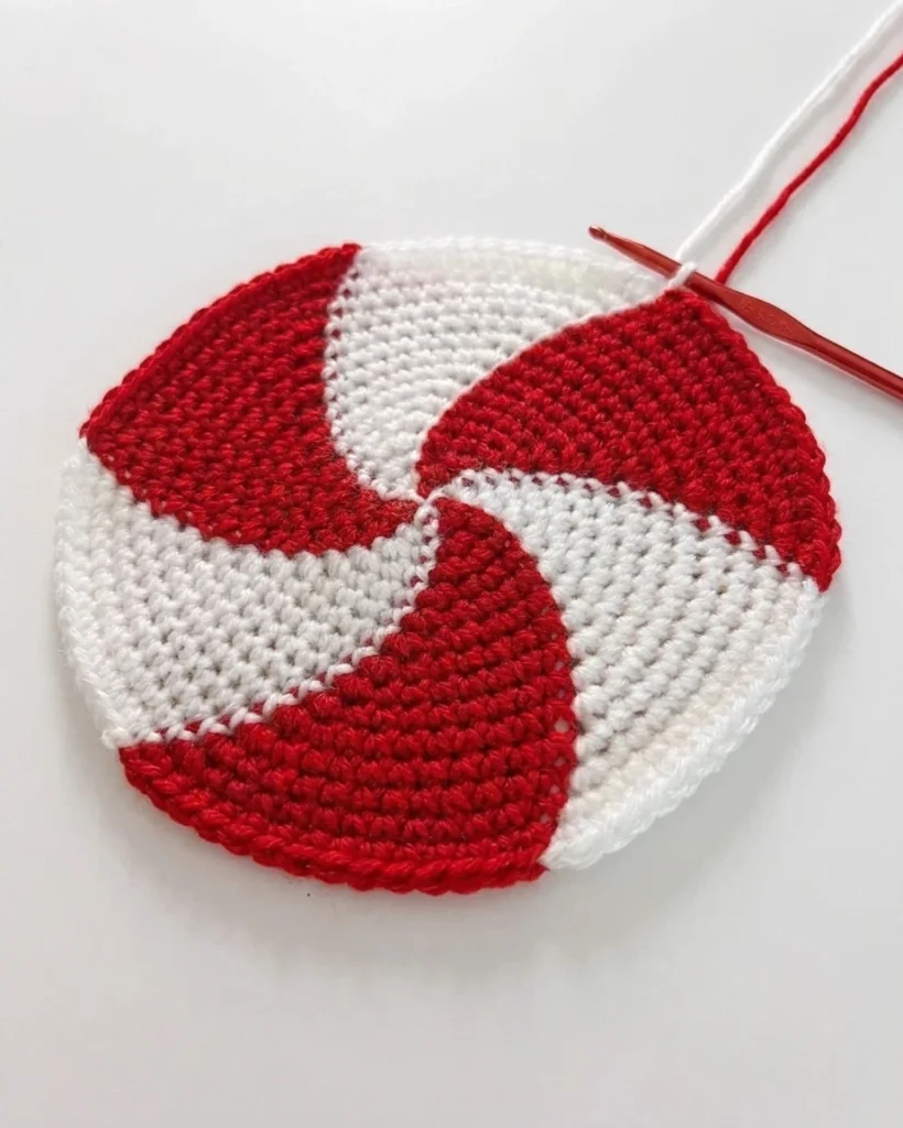 A red-and-white crocheted spiral hat crown with a hook and working yarn attached.