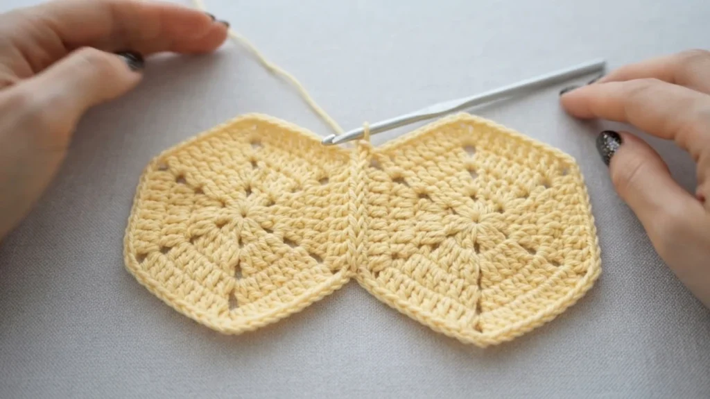 Two pale yellow crochet hexagons joined edge to edge, a crochet hook resting in the seam, with hands holding the work.