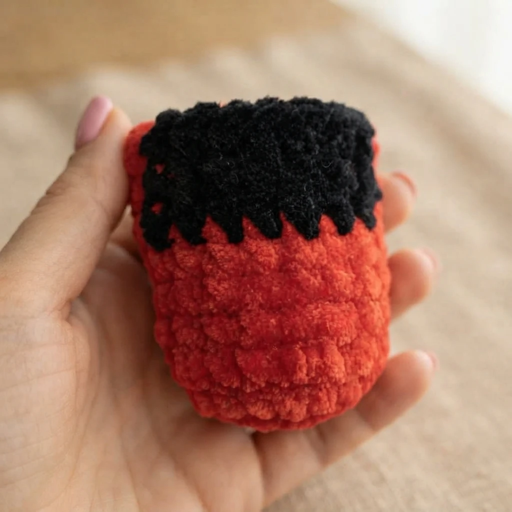 A hand holds an unfinished red crocheted bird body with a black section across the top.