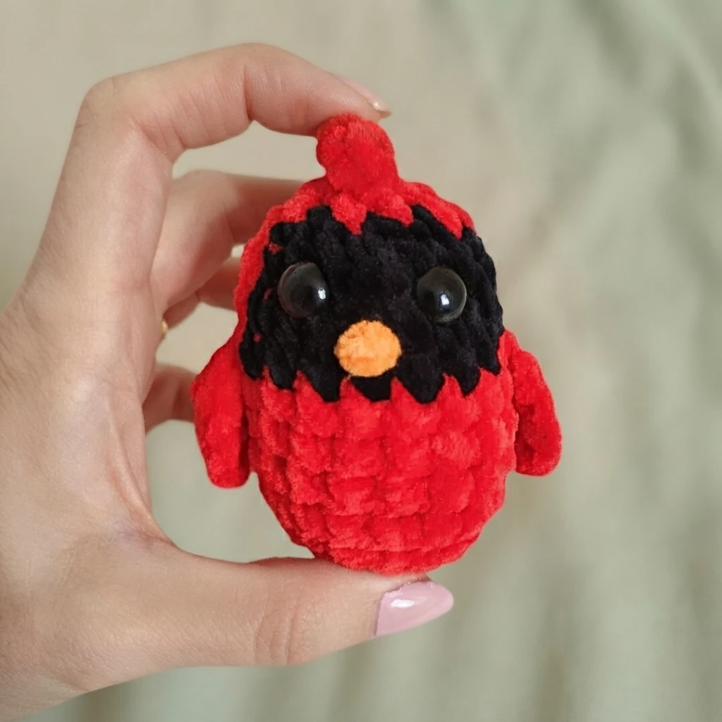 A hand holds a red crocheted cardinal with a black face, orange beak, and plastic eyes.
