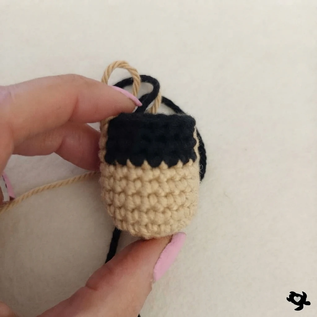 A hand holds an unfinished beige crochet bird body with a black upper section and loose yarn strands.