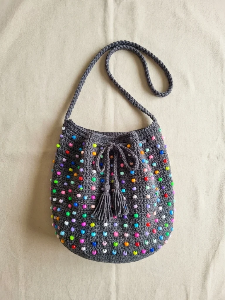 Grey crocheted beaded bucket bag with colorful beads, drawstring closure and long strap, laid flat on cream fabric.