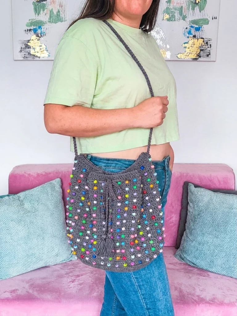 A woman wearing a grey crocheted bucket bag with colorful beads over her shoulder in a living room