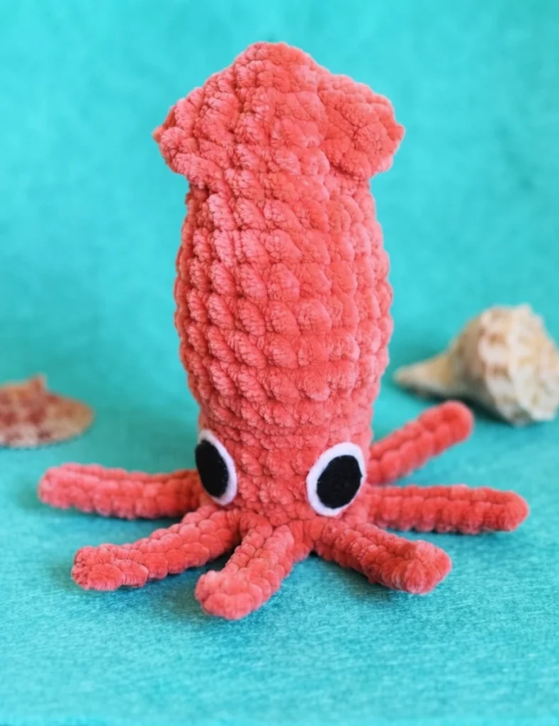 Coral chenille crochet squid amigurumi with large black and white felt eyes and eight tentacles, posed on a turquoise background with small seashells.