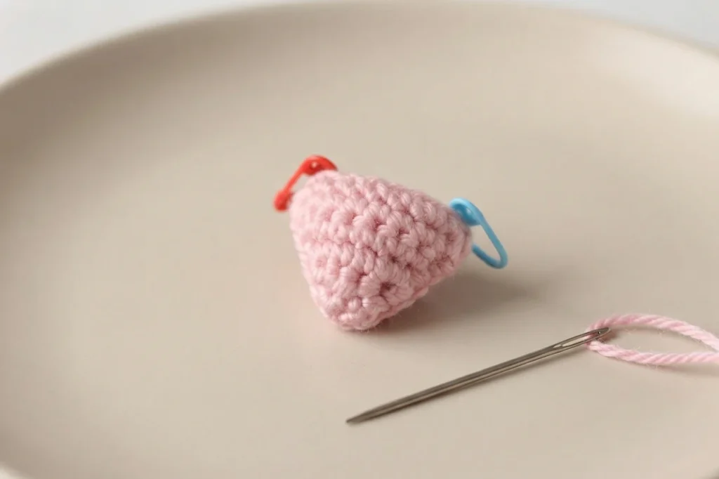 Small pink crocheted triangular nose piece with a red and a blue stitch marker, plus a threaded yarn needle, resting on a plain beige surface.