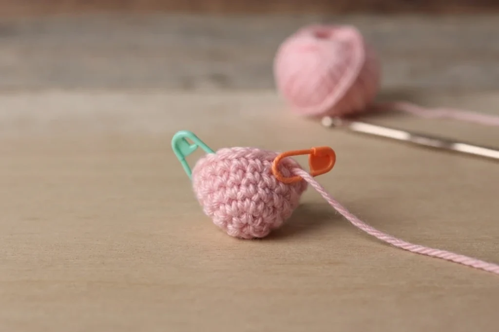 Pink crocheted nose piece resting on a wooden surface, with a teal stitch marker on the left and an orange stitch marker on the right, and a blurred ball of pink yarn and metal crochet hook behind it.