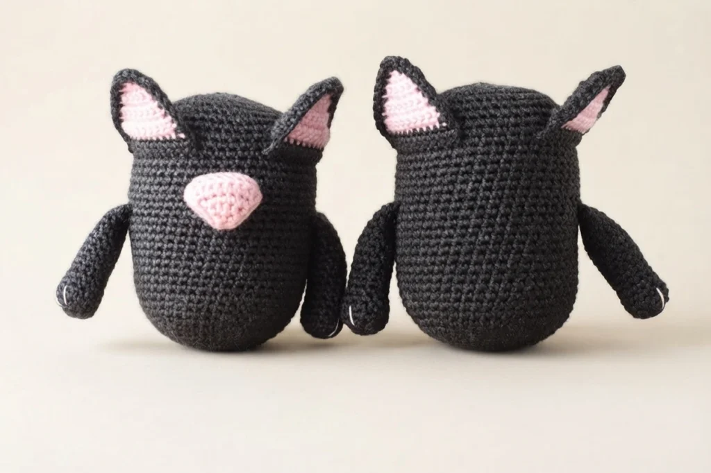 Two crocheted cat gnome bodies in dark charcoal yarn, each with pink-lined ears and a pink muzzle, and small arms attached at the sides; one body shows a pink nose patch on the front while the other is shown from behind.
