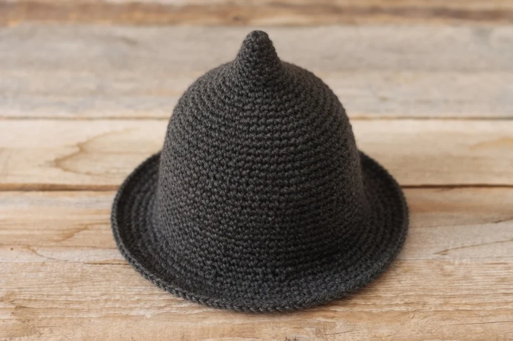 Crocheted dark grey hat with a rounded crown, a short point on top and a wide flat brim, resting on a light wooden surface.