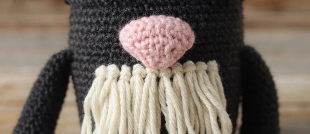 Close-up of a crocheted cat gnome showing a pink stuffed nose with cream yarn loops attached underneath in a row to form a beard, set against a dark charcoal grey crocheted body with the tops of the arms visible at the sides.