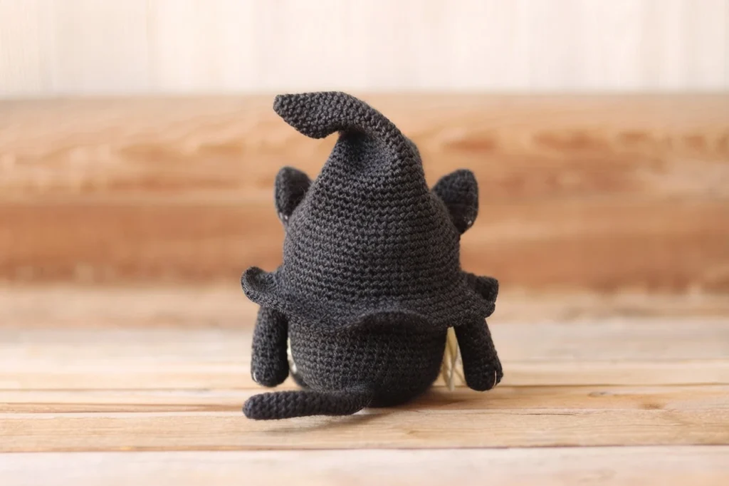 Back view of a dark charcoal crochet cat gnome with a pointed witch hat, small ears, short arms, a curled tail and a few light strands showing at the side, sitting on a wooden surface.