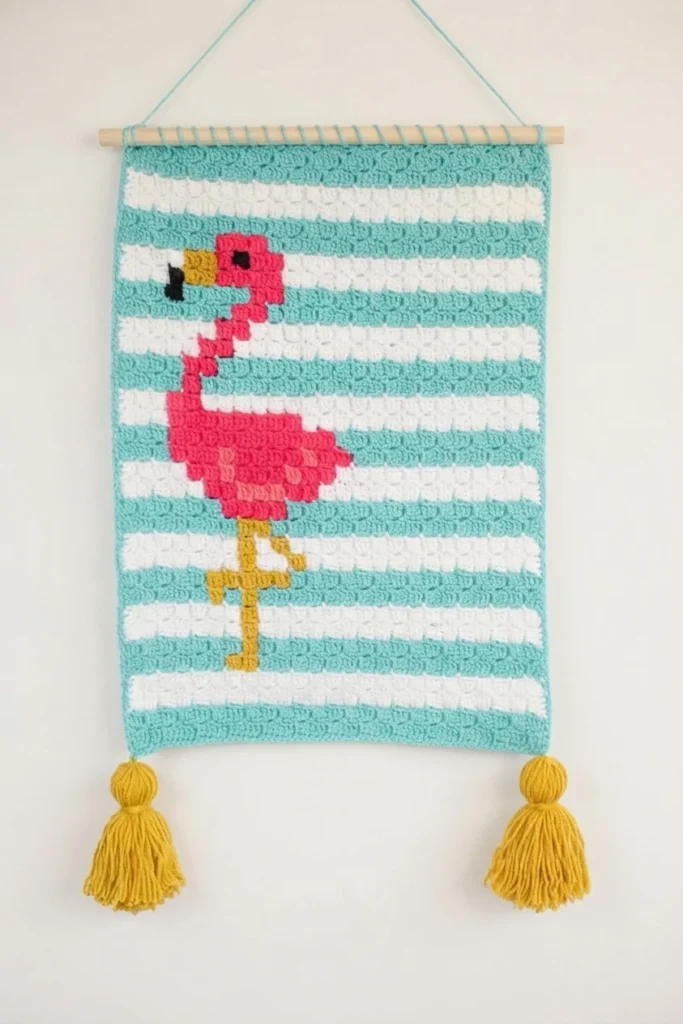 Finished corner to corner crochet wallhanging with teal and white stripes, a pink flamingo, two gold tassels and a wooden dowel hanging from a cord