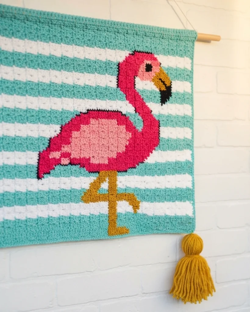 Crocheted corner to corner wallhanging hanging from a wooden dowel, showing a pink flamingo with light pink wing, gold legs and black outline on teal and white stripes, with a mustard yellow tassel at the bottom.