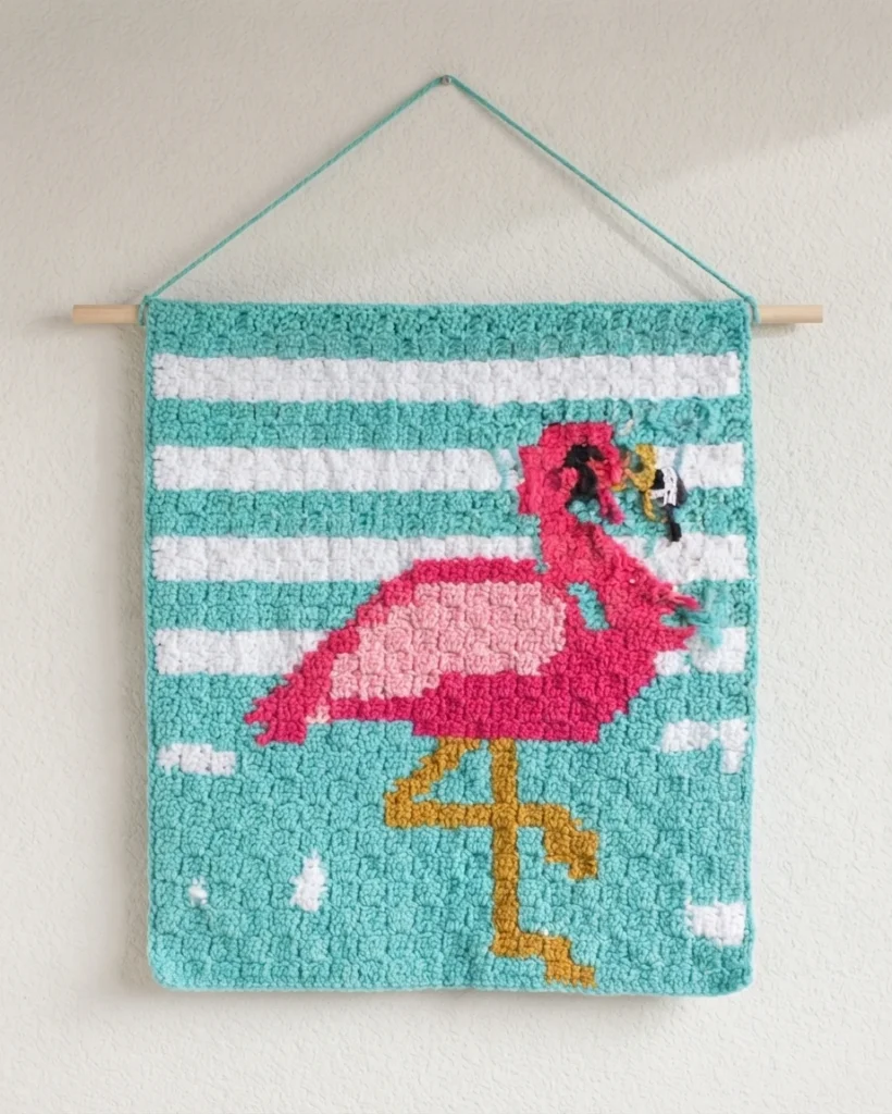 Finished corner to corner crochet flamingo wallhanging in teal, white, pink and gold stripes hanging from a wooden dowel on a wall, with a few loose yarn ends still visible near the flamingo's head