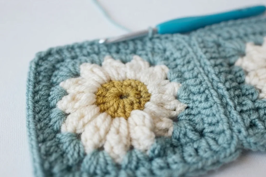 Close-up of a crocheted daisy granny square with cream petals, a yellow center and a teal background, with a blue-handled crochet hook resting at the edge of the square.