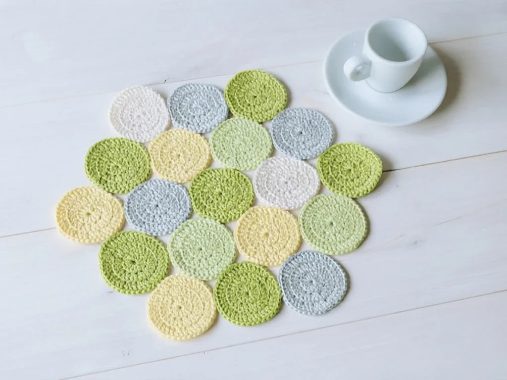 Crocheted placemat made of small joined circles in pale green, yellow, white and light grey, arranged in a hexagonal shape on white painted wooden boards, with a white cup on a saucer at the upper right.