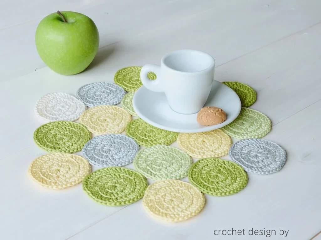 Crocheted placemat made of overlapping round motifs in green, white, pale yellow and grey on a white wooden table, with a green apple at the upper left and a white cup on a saucer holding a small biscuit.