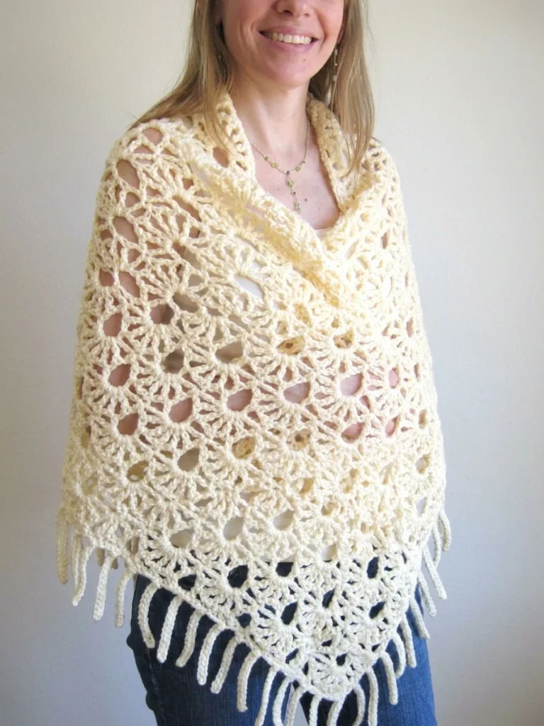 Person wearing a cream lacy crochet shawl with openwork fan motifs and long fringe, wrapped and crossed over the shoulders above blue jeans.