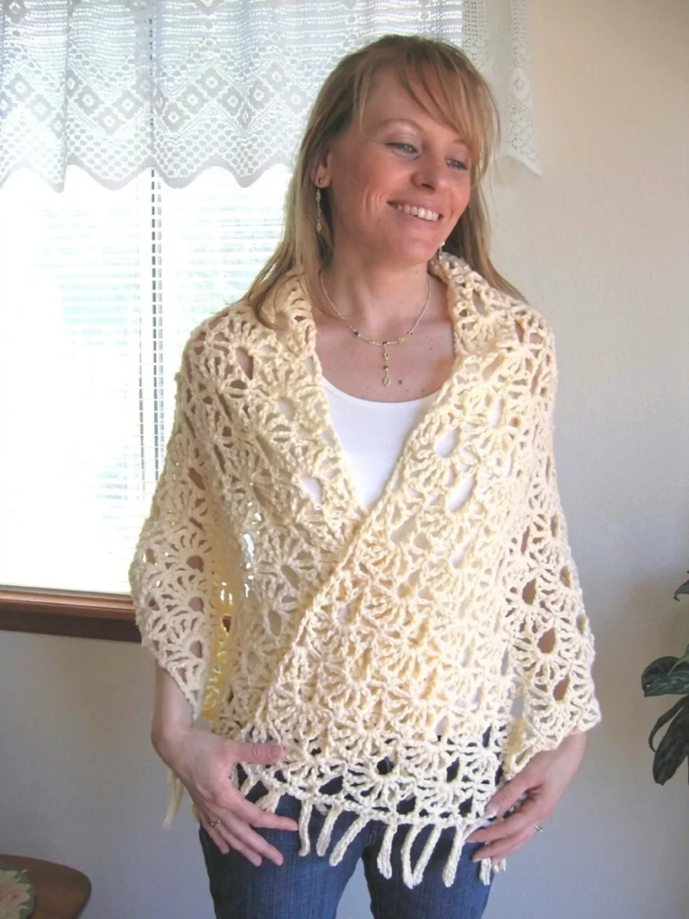 Woman smiling in a cream lacy crocheted shawl with open fan motifs and long fringe, wrapped diagonally over a white top and blue jeans, standing beside a window with a white lace curtain and a potted plant at right.