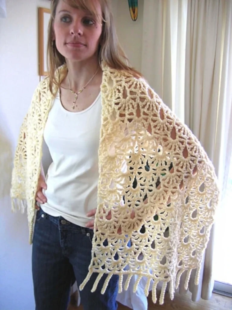 A woman standing indoors wears a cream openwork crochet shawl with large fan and diamond motifs and long chain fringe, holding the shawl edges open over a white top and blue jeans.