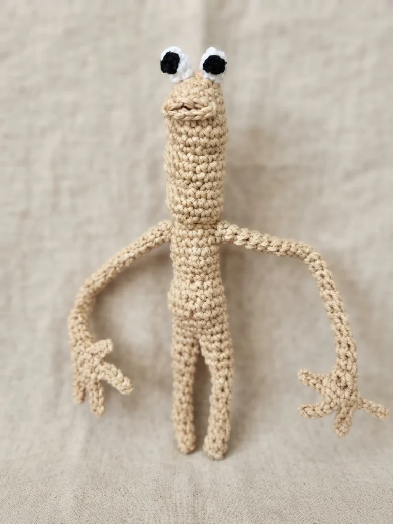 Crocheted beige worm figure with crocheted white and black eyes and a small stitched mouth, two long thin arms ending in three-fingered hands and two straight legs, standing upright on a light beige linen fabric.