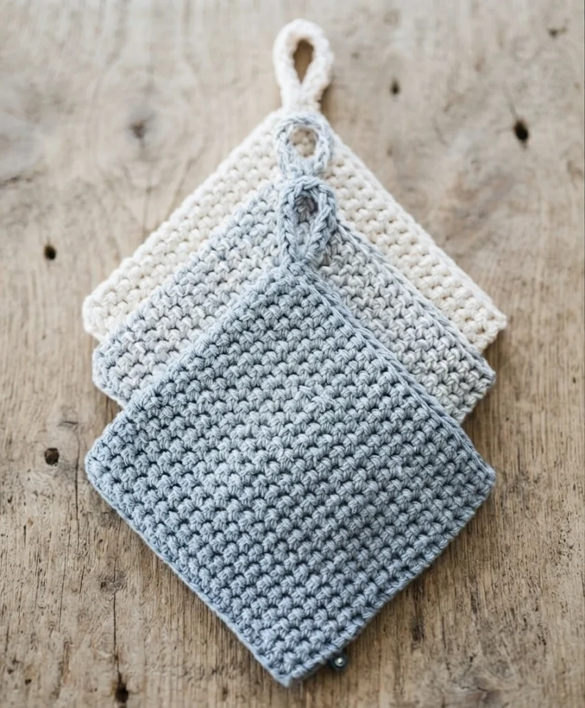 Three stacked square crocheted potholders in cream and light grey with looped hanging tabs, resting on a rustic wooden surface.
