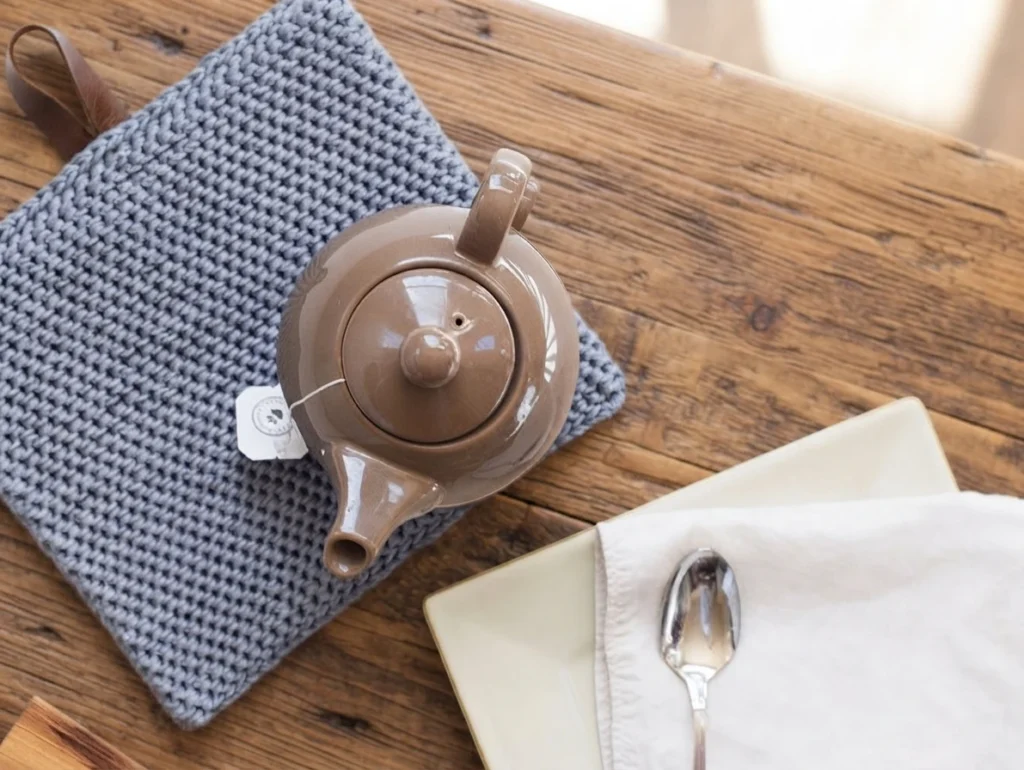 Gray textured crochet potholder with a brown leather hanging loop, holding a glossy brown ceramic teapot, next to a white plate, a folded napkin and a spoon on a wooden table.