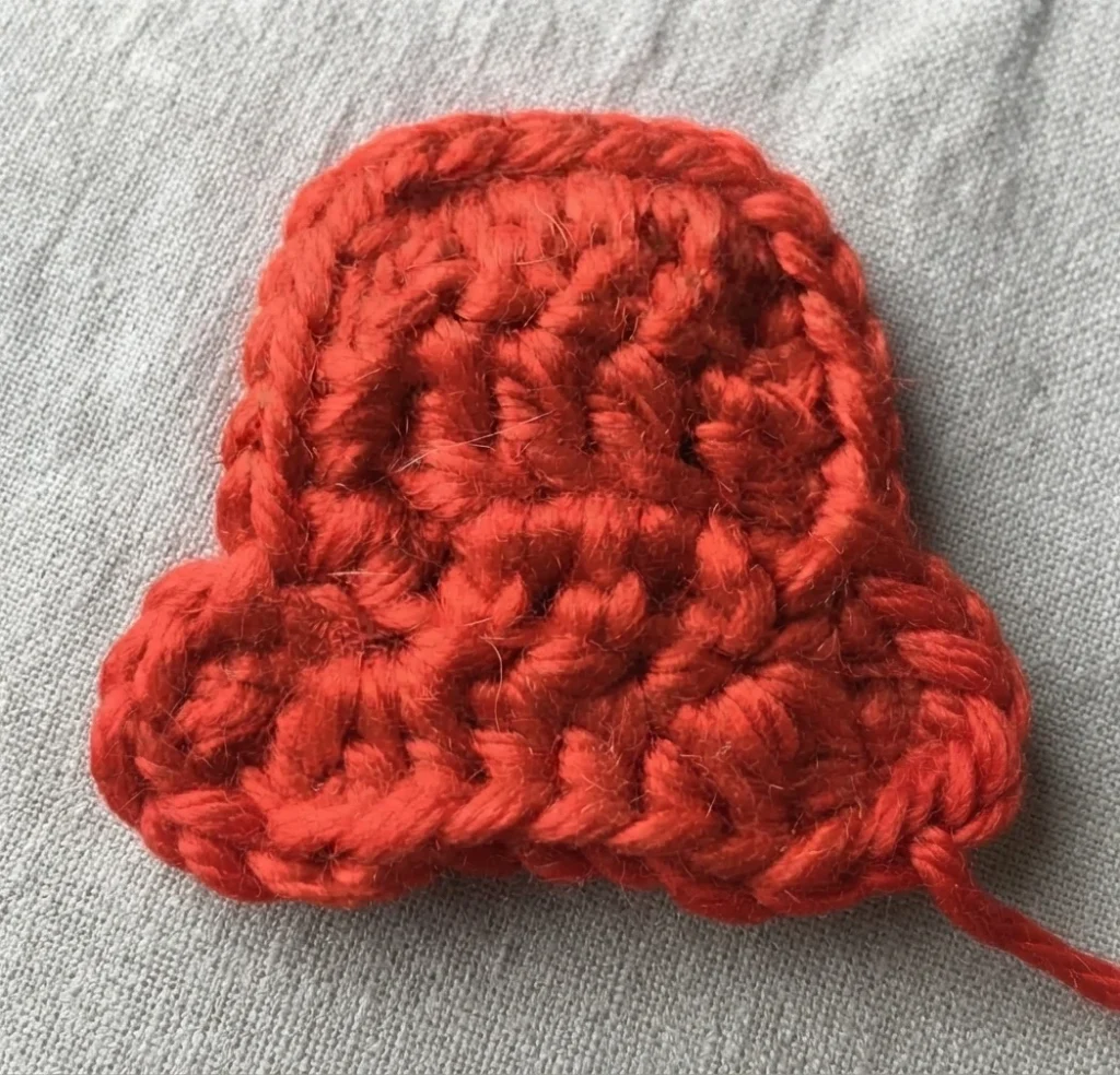 Crochet red top hat piece worked in double crochet with a neatened single crochet edge, shown alone on a light grey fabric background.
