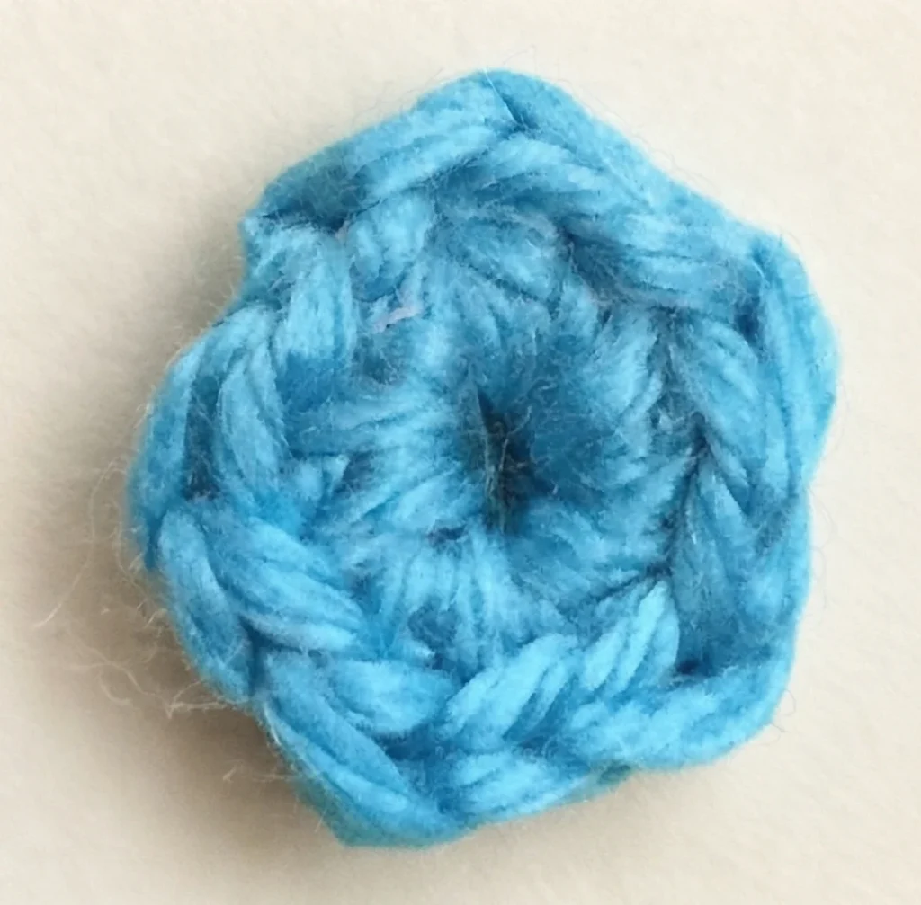 Close-up of a small blue crocheted flower motif with five rounded petals and a tight center, resting on a plain light beige surface.