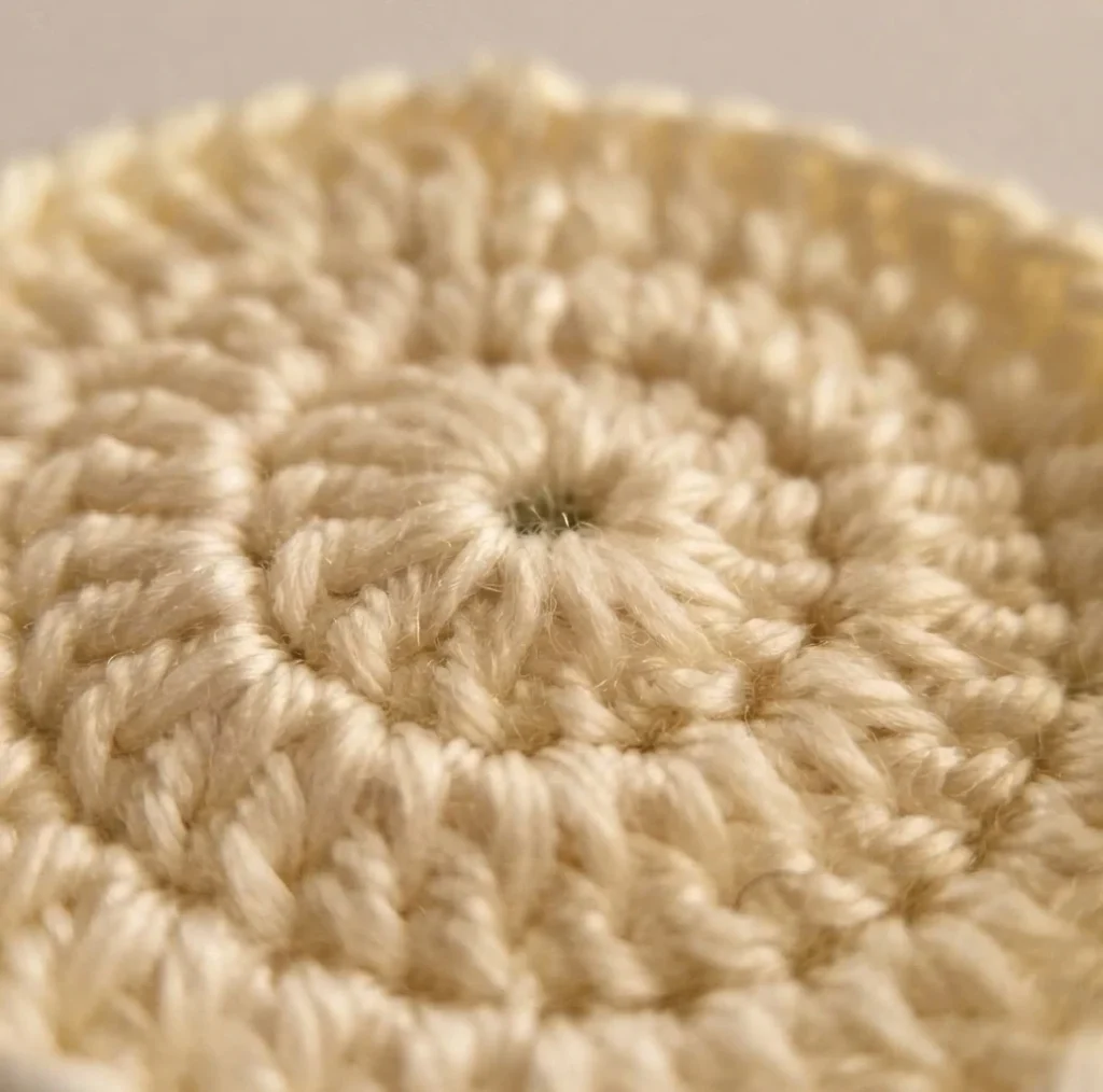 Close-up of a cream crocheted circle worked in concentric double crochet rounds around a small dark center, photographed at an angle with a blurred neutral background.