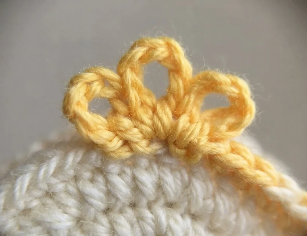 Close-up of pale yellow crocheted chain loops forming the hair along the top edge of a cream crocheted head piece, with a loose yarn end trailing to the right.
