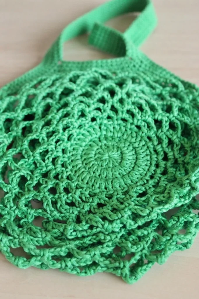 Flat lay of an emerald green crochet mesh bag with a solid round base, open netted body and crocheted strap, on a light wooden surface.