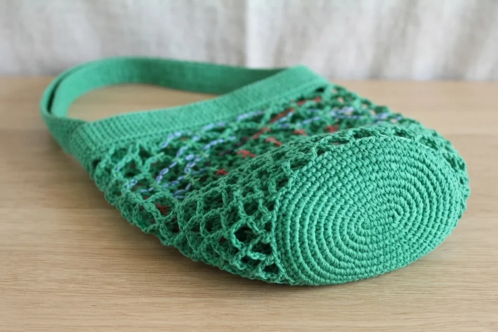 Emerald green crocheted mesh market bag with a solid round base and wide strap, lying on its side on a wooden table