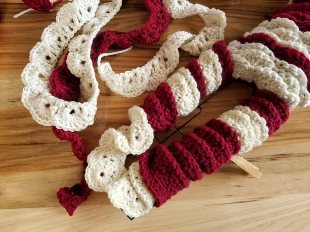 Crocheted burgundy and cream strips with a scalloped shell texture lying on a wood plank floor beside a black wire candy cane shaped wreath frame, with one strip attached to the frame by a wooden clothespin