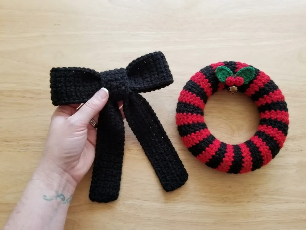 Hand holding a large black crocheted bow above a light wood table, next to a small circular wreath with red and black stripes, green holly leaves, red berries and a tiny bell.