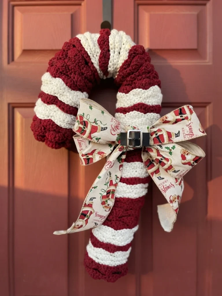 Crocheted candy cane wreath in burgundy and cream stripes hanging on a red door, decorated with a Santa print ribbon bow and a black belt with a silver buckle at the crook