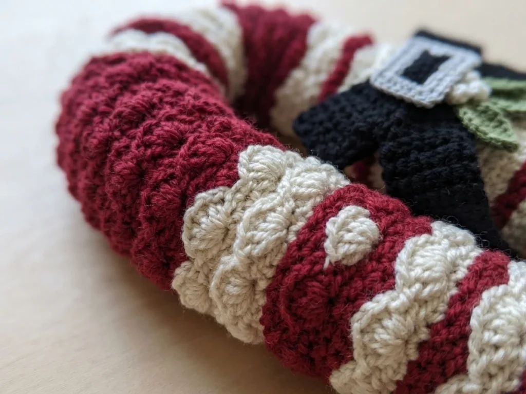 Close-up of a crocheted candy cane wreath worked in deep red and cream shell stitch stripes, with a black crocheted bow, a silver buckle and small green mistletoe leaves, photographed against a softly blurred pale surface.