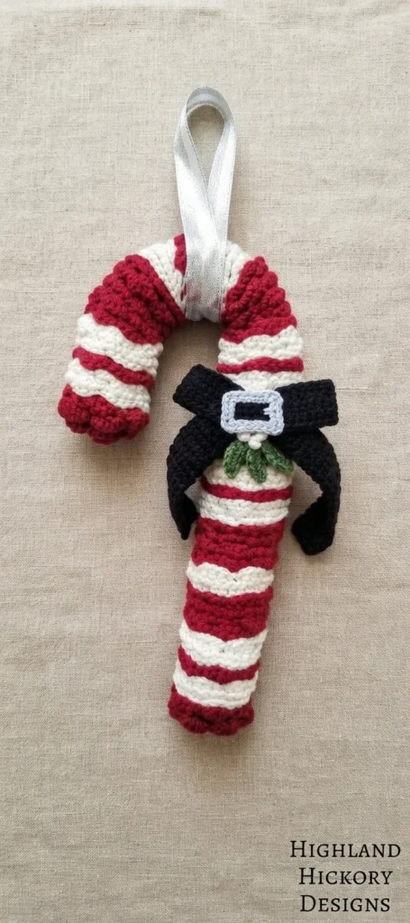 Crocheted candy cane wreath in red and white stripes with a silver ribbon loop at the top, a black bow with a silver buckle and green mistletoe at the curve, photographed on beige fabric.