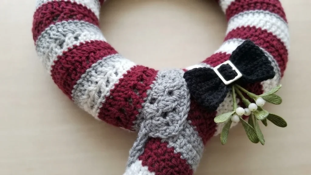Close-up of a crocheted wreath with alternating burgundy, white and light grey striped sections, a black crochet bow with a silver buckle and a sprig of faux mistletoe with white berries.