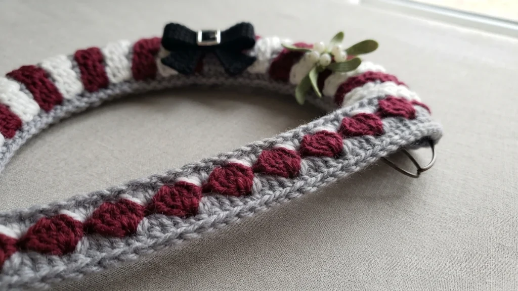 Close-up of a crocheted wreath section with burgundy and white stripes worked over a grey base, decorated with a small black bow with a silver buckle and a sprig of mistletoe with white berries, resting on a pale fabric surface with a metal
