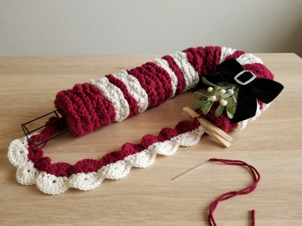 Candy cane wreath in progress on a light wooden table: a partly wrapped candy cane wire frame with burgundy and white striped crochet strips, a loose ruffled strip laid in front, a wooden clothespin, a tapestry needle threaded with burgundy