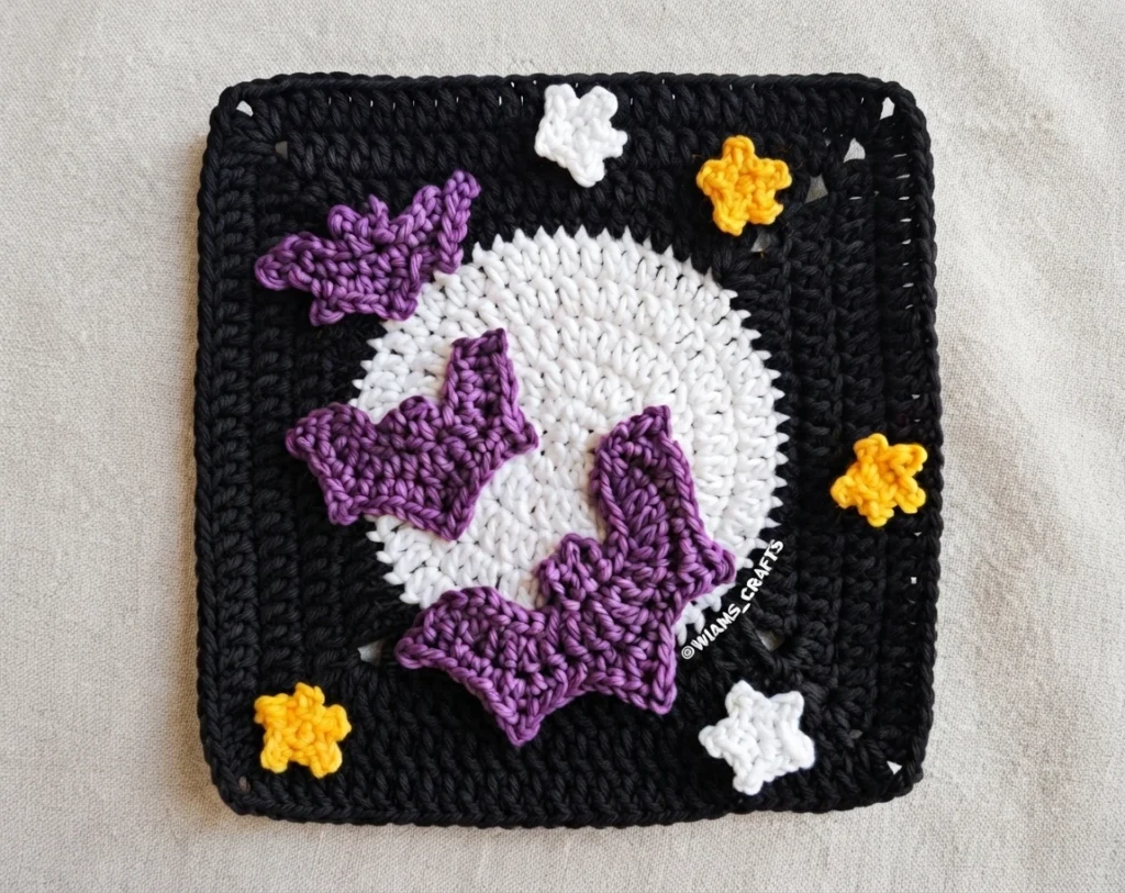 Carré au crochet noir décoré d’une lune blanche, de trois chauves-souris violettes et d’étoiles jaunes et blanches.