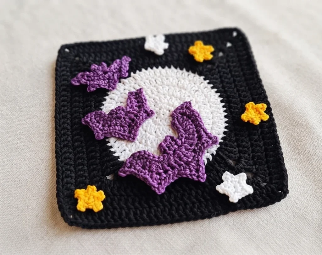 A black crocheted square with a large white moon, three purple bats, and small white and yellow motifs on a pale surface.