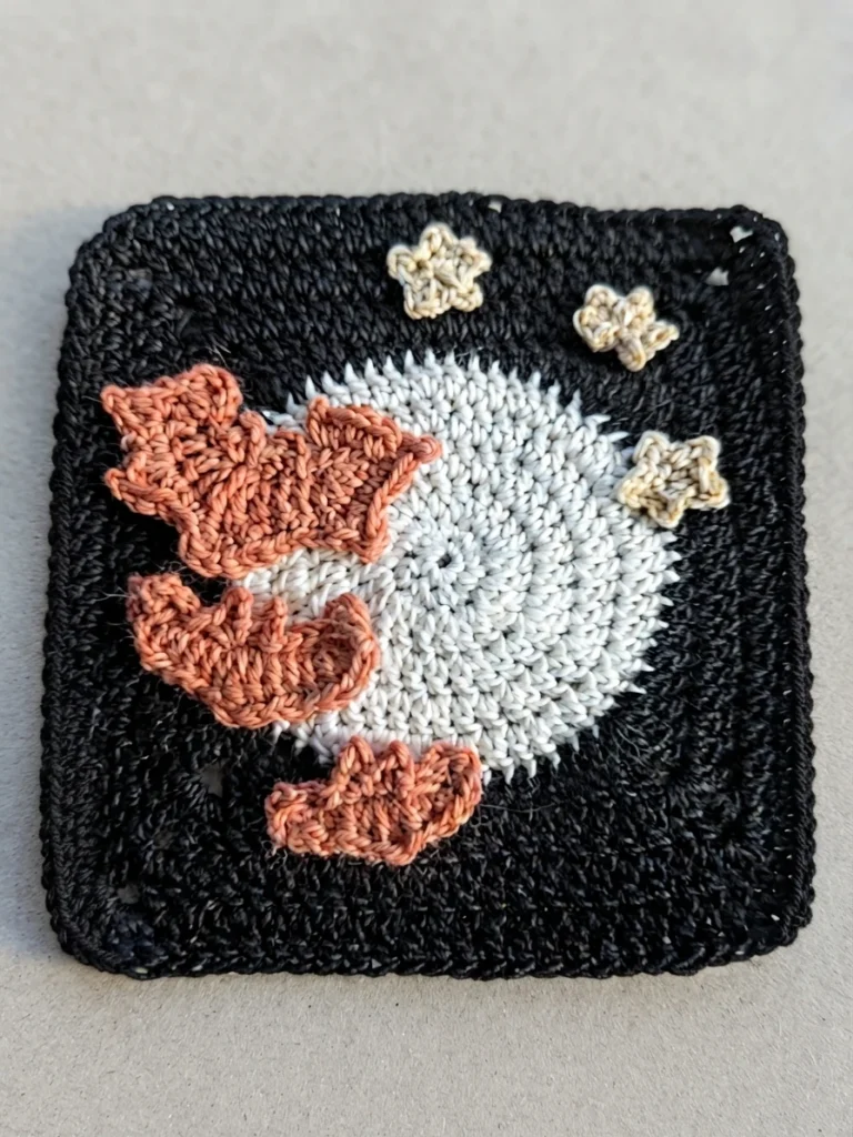 Black crocheted square with a white circular center, three orange bat appliqués, and three beige stars.