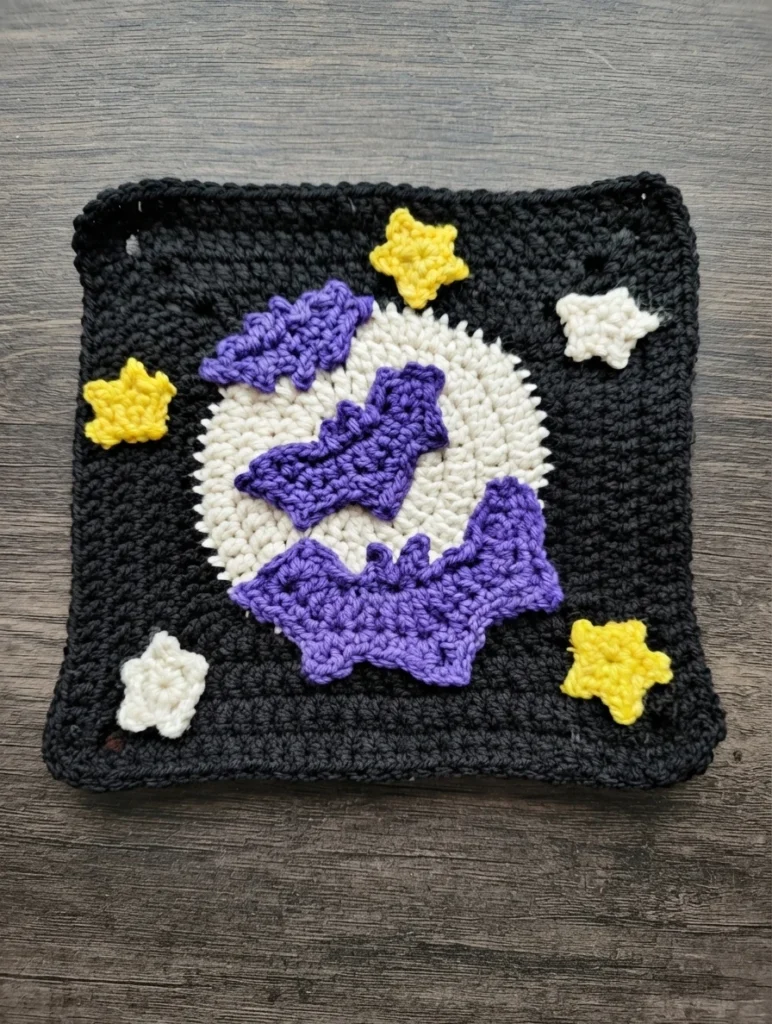 A black crocheted square with a white moon, three purple bats, and yellow and white stars, photographed on a wooden surface.
