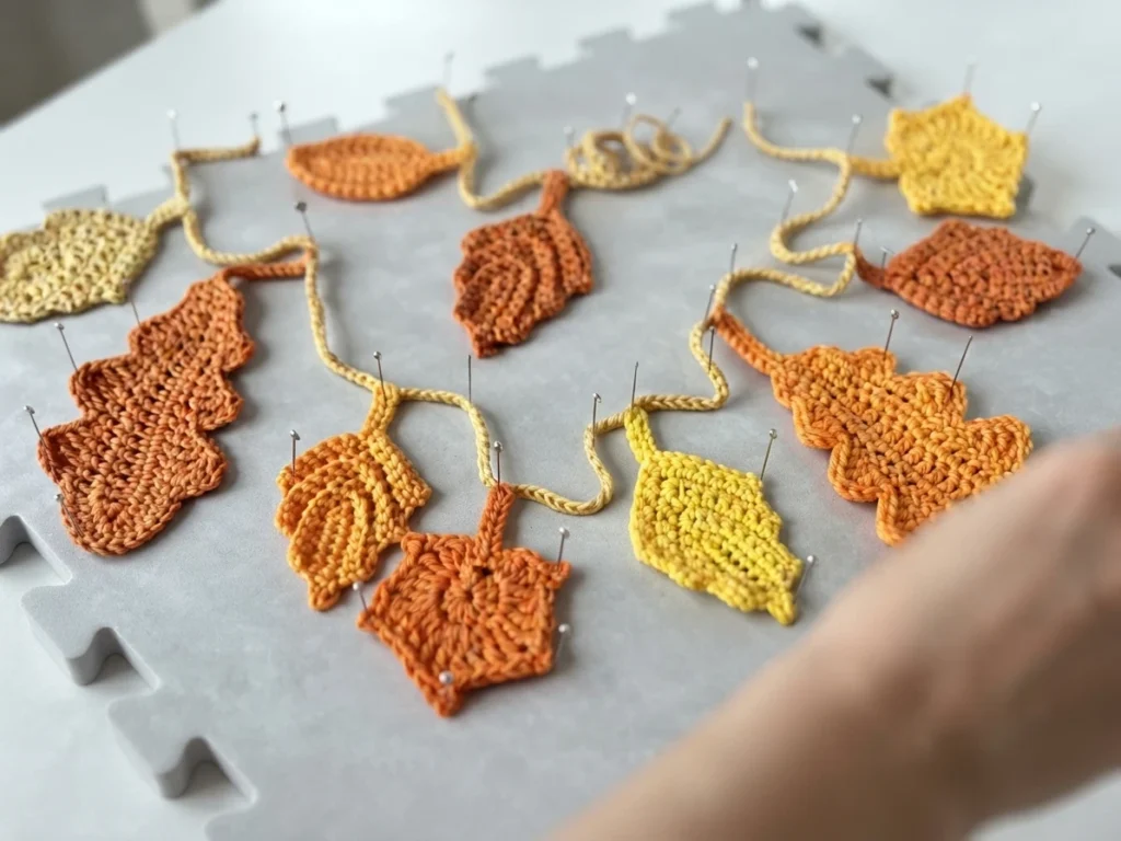 Crochet autumn leaves in yellow, orange and rust pinned to a gray blocking mat along a pale yellow chain