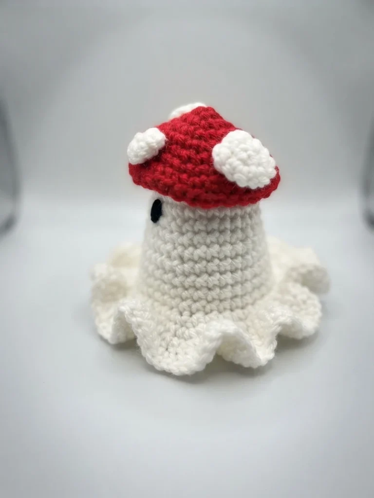 Crocheted ghost amigurumi with a red cap dotted with white spots, a black oval safety eye and a ruffled white skirt, photographed against a plain white background.