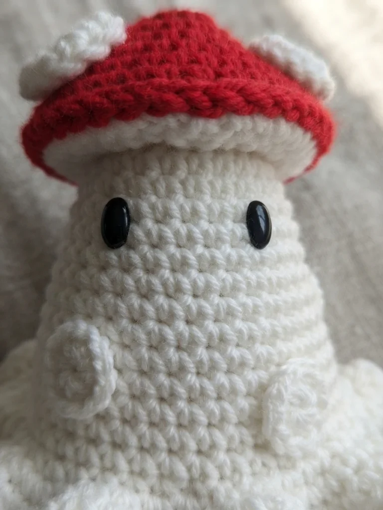 Close-up of a crocheted toy with a red cap dotted with white spots, a white crocheted body, two black oval safety eyes and a ruffled white base, resting on light grey fabric.