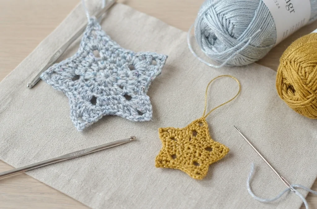 Two sparkly crocheted five-point stars in silver and gold beside yarn balls, crochet hooks and a yarn needle.