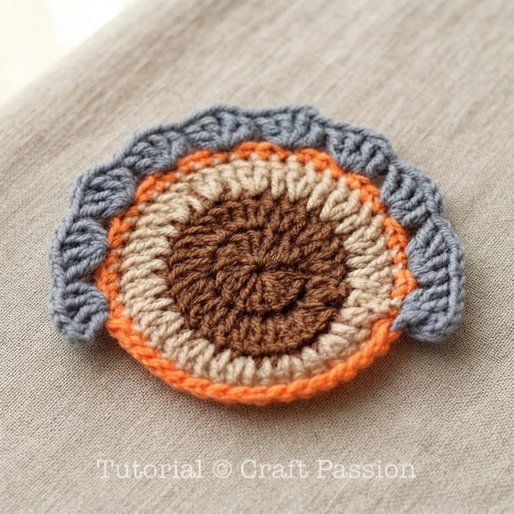 Crochet turkey coaster in progress: a round piece with a dark brown center, a cream ring, an orange outer ring and a grey scalloped tail edge, laid on plain beige woven fabric with a Tutorial © Craft Passion watermark.