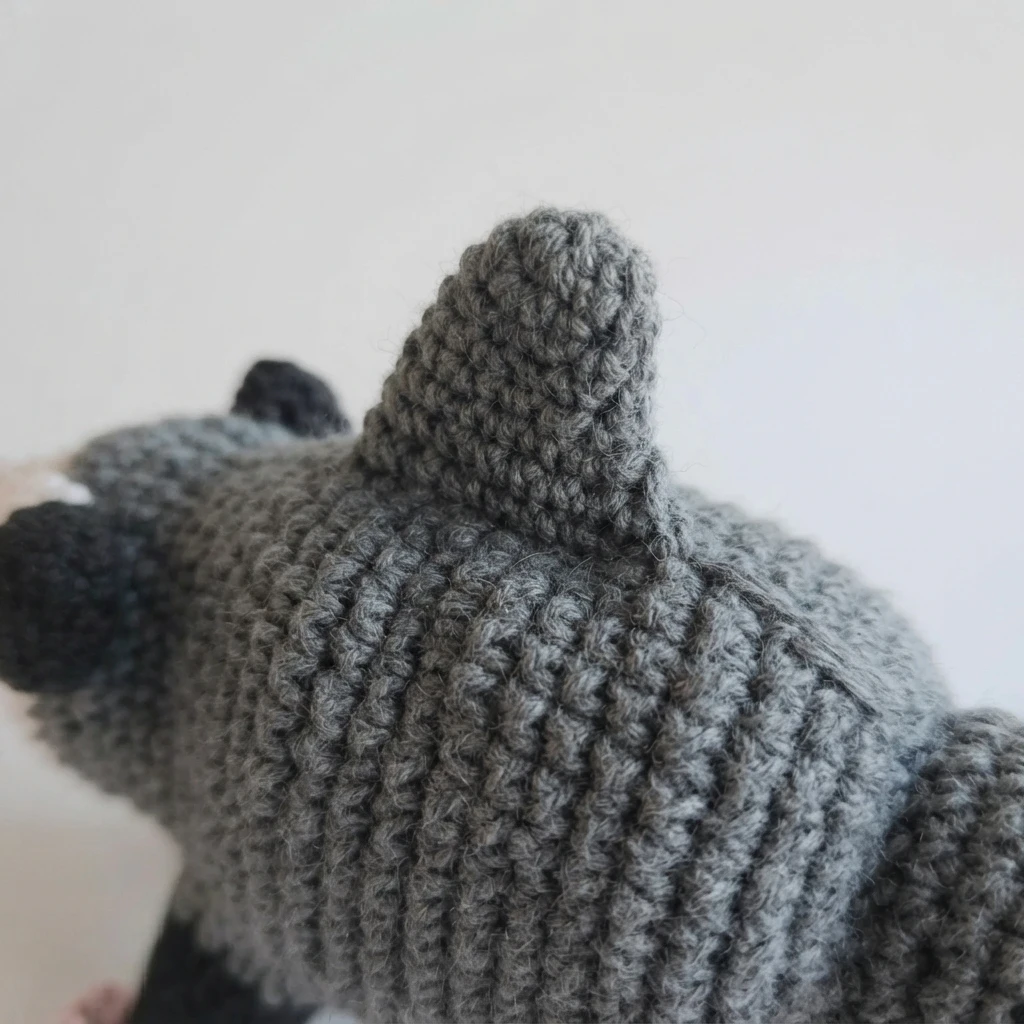 Close-up of a crocheted gray shark possum from behind, showing the large triangular back fin sewn along the spine and a short loose yarn tail at the base of the fin.